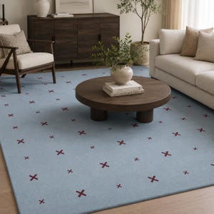 May include: A light blue area rug featuring a repeating pattern of small, dark red cross symbols distributed evenly across the surface. The rug has a low-pile texture and covers the floor space beneath a round wooden coffee table and furniture.
