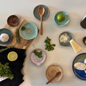 Peut inclure: Une collection d'ustensiles de cuisine en céramique comprenant plusieurs petites assiettes à râper rondes dans des tons bleu, vert et rose, accompagnées de cuillères en bois, d'une planche à découper, d'herbes fraîches, de citron vert, d'ail, de fromage et de pain tranché sur une surface blanche.
