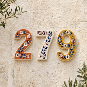 May include: Three ceramic house numbers, 2, 7, and 9, featuring hand-painted floral patterns. The number 2 is orange, the number 7 is cream, and the number 9 is yellow. Each digit is decorated with blue vines, white flowers, and red tulip buds.