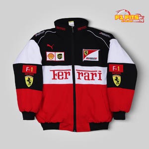 Könnte beinhalten: Eine schwarz-weiß-rote Reißverschlussjacke mit dem Ferrari-Logo, dem Schriftzug Ferrari in großen roten Buchstaben und verschiedenen Sponsoren-Aufnähern wie Shell, UPS und Bridgestone. Die Jacke hat einen Stehkragen und lange Ärmel mit F-1-Aufnähern.