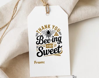Puede incluir: Una etiqueta de regalo rectangular blanca con un cordel de yute marrón. La etiqueta presenta un gráfico en negro y dorado de una abeja, un panal y el texto THANK YOU BEE-ING FOR SWEET y From, impreso en una fuente decorativa y llamativa.