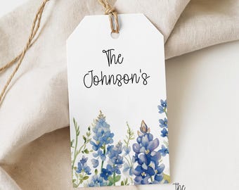 Puede incluir: Una etiqueta de regalo rectangular blanca con esquinas superiores redondeadas, que presenta una ilustración en acuarela de flores de lupino azules en la parte inferior y el texto negro The Johnson's impreso en el centro, unida a un cordel de yute marrón.