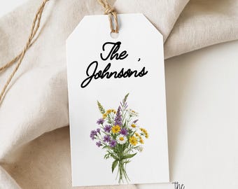Puede incluir: Una etiqueta de regalo rectangular blanca con un agujero en la parte superior y un cordel marrón. La etiqueta presenta el texto negro The Johnson's y una ilustración colorida de un ramo de flores silvestres con flores amarillas, moradas y blancas.