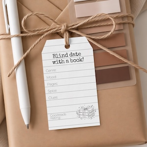 Könnte beinhalten: Ein weißes rechteckiges Papieretikett mit schwarzen horizontalen Linien und dem Text Blind date with a book!, befestigt mit brauner Schnur an einem braunen Paket. Ein weißer Stift und eine transparente Kunststofffolie mit fünf braunen Klebestreifen.