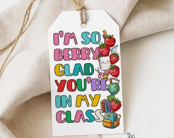 Puede incluir: Una etiqueta de regalo rectangular blanca con un cordel marrón que presenta el texto colorido I'M SO BERRY GLAD YOU'RE IN MY CLASS. El diseño incluye ilustraciones de fresas sonrientes, un cuaderno, tijeras, un globo terráqueo y una mochila.