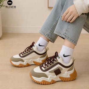Può includere: Un paio di sneakers spesse con tomaia color crema e verde oliva, lacci e finiture marrone scuro. Le scarpe presentano una suola in gomma spessa e ondulata color gomma con battistrada testurizzato. Si indossano calzini bianchi con logo nero.