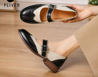May include: A two-tone Mary Jane shoe featuring a black toe cap and heel counter with decorative perforations, contrasted by a cream-coloured side panel. The shoe includes a black adjustable strap with a silver-toned metal buckle and a low brown heel.