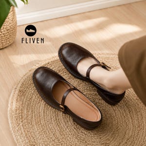 May include: A pair of dark brown leather Mary Jane shoes featuring a rounded toe, a thin adjustable strap across the instep with a small gold-toned buckle, and a low heel. The brand name FLIVEN is printed in black text above the shoes.