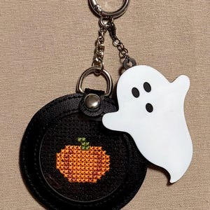 May include: A circular black keychain featuring a cross-stitched orange pumpkin with a green stem in the center. Attached to the silver metal ring and chain is a flat, white, ghost-shaped charm with two black oval eyes and a black oval mouth.