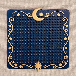 May include: A square dark blue grid panel featuring a raised gold crescent moon at the top center and a gold eight-pointed star at the bottom center. The border is decorated with gold swirling filigree lines and small gold stars.