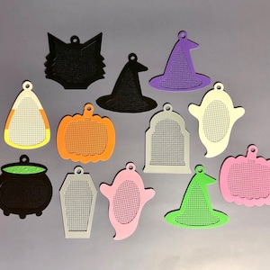 May include: A collection of twelve assorted Halloween-themed plastic pendants featuring a perforated grid center. Shapes include a black cat head, witch hats in black, purple, and green, pumpkins in orange and pink, ghosts, a cauldron, and tombstones.
