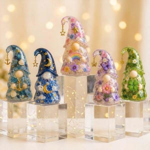 Puede incluir: Cinco figuras de gnomos de resina con elementos florales, celestiales y náuticos incrustados. Cada gnomo tiene un sombrero puntiagudo con un dije colgante, una nariz redonda y sostiene un objeto específico como un ancla, luna, arcoíris, mariposa o trébol.