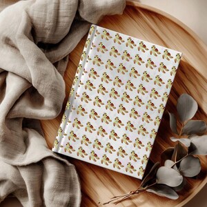 May include: A hardcover journal featuring a repeating pattern of small, cartoon-style sea turtles with green and yellow shells on a white background. The notebook has a clean, rectangular shape with a smooth, matte-finish cover design.