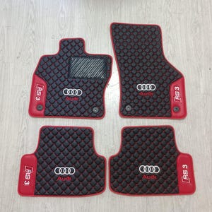 Könnte beinhalten: Ein Set aus vier schwarzen Autofußmatten mit rotem Rautensteppmuster und roter Umrandung. Jede Matte zeigt das weiße Audi-Logo und das rote RS 3-Emblem. Die Fahrermatte verfügt über eine strukturierte schwarze Fersenauflage und zwei Befestigungslöcher.
