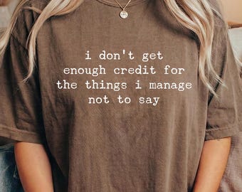May include: A brown short-sleeve t-shirt featuring the white printed text: i don't get enough credit for the things i manage not to say. The shirt has a crew neckline and a relaxed fit, made from a solid-colored fabric with a subtle textured appearance.