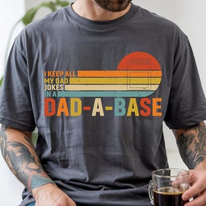 May include: A charcoal grey short-sleeve t-shirt featuring a retro-style graphic with horizontal stripes in orange, yellow, and light blue. The text reads I KEEP ALL MY DAD JOKES IN A DAD-A-BASE, accompanied by a small icon of a memory card.