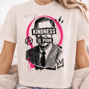May include: A cream-colored short-sleeve t-shirt featuring a black and white graphic of a man in a sweater and tie. The design includes a bright pink circle, an anarchy symbol, and the text "KINDNESS IS PUNK" printed over the eyes and mouth.