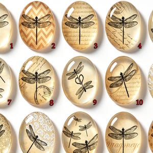 Op de afbeelding: Een collectie van vierentwintig ovale glazen cabochons met zwarte libelle-illustraties. Elk stuk toont unieke achtergrondpatronen, waaronder vintage schrift, chevron, stippen, bloemmotieven en hartjes, genummerd van één tot vierentwintig.