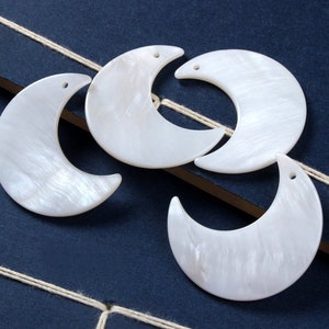 May include: Four white crescent moon shaped shell charms with holes for hanging. The charms are arranged in a pile on a blue surface.