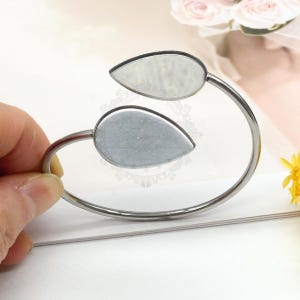 Puede incluir: Una pulsera de puño plateada con dos engastes en forma de lágrima. La pulsera está sujeta por una mano, y los engastes parecen estar diseñados para sujetar una piedra u otro elemento decorativo. La pulsera tiene un diseño sencillo y elegante.