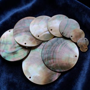 May include: A collection of iridescent, round mother-of-pearl shell discs in various sizes. Each disc has a small hole near the edge. The shells display a mix of colors, including green, pink, and gold, with a natural, swirling pattern.