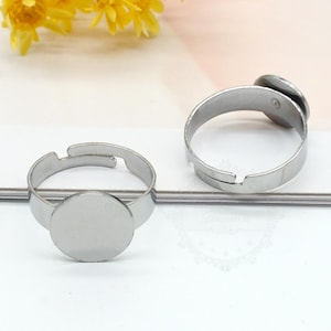 Könnte beinhalten: Zwei silberfarbene, verstellbare Ringe mit runden, flachen Fassungen. Die Ringe bestehen aus glänzendem Metall und sind für die Aufnahme einer kleinen Verzierung oder eines Cabochons konzipiert. Sie werden auf einer weißen Oberfläche mit gelben Blumen im Hintergrund präsentiert.