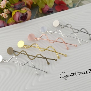Pode incluir: Uma coleção de grampos de cabelo decorativos em vários acabamentos metálicos, incluindo prata, ouro e cobre. Cada grampo apresenta um design ondulado com um acento circular e uma pequena bola na extremidade. Os grampos estão dispostos sobre uma superfície branca.