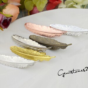 Puede incluir: Cinco abalorios de metal con forma de pluma en diferentes colores: plata, oro, oro rosa, latón antiguo y cobre. Los abalorios están dispuestos en fila sobre una superficie blanca. Gemstones2u está escrito en texto negro en la parte inferior de la imagen.