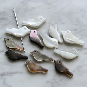 May include: A collection of bird-shaped beads in various colors, including white, black, and iridescent shades. A silver needle is also present. These beads are likely for jewelry making or crafting.