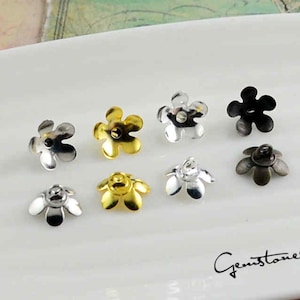 Może przedstawiać: Kolekcja elementów biżuteryjnych w kształcie kwiatów w różnych metalicznych wykończeniach, w tym srebrnym, złotym i czarnym. Elementy są ułożone na białej powierzchni, prezentując szczegółowe wzory płatków. Obraz zawiera tekst "Gemstones2u".