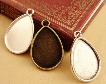 May include: Three teardrop-shaped pendant trays with silver and bronze finishes. The trays are designed to hold a small piece of glass or other material for jewellery making.
