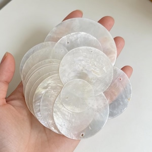 Puede incluir: Un conjunto de discos redondos e iridiscentes de concha de nácar. Cada disco tiene un pequeño agujero cerca del borde. Los discos, de diferentes tamaños, se sostienen en una mano, mostrando su brillo nacarado natural. Ideal para manualidades o joyería.