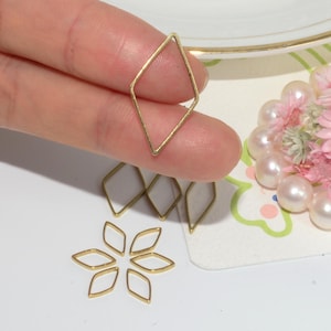 Pode incluir: Close-up de vários pequenos pingentes de metal dourado em forma de diamante. Um é segurado entre os dedos, enquanto outros estão espalhados abaixo. Alguns pingentes são dispostos em forma de flor. Pérolas e flores cor-de-rosa ao fundo.