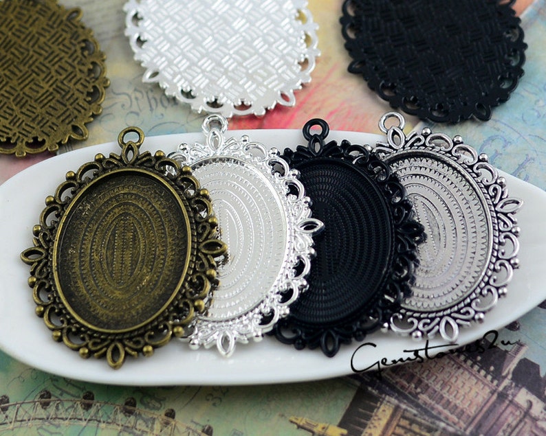Puede incluir: Una colecci&oacute;n de bandejas colgantes ovaladas con bordes de filigrana ornamentados. El conjunto incluye cuatro engastes de metal en acabados bronce antiguo, plata y negro, cada uno con un &aacute;rea central rebajada y una anilla superior.