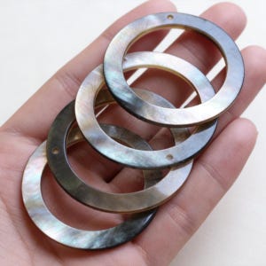 May include: Four iridescent, circular mother-of-pearl jewellery components. Each ring has a small hole and a dark grey outer edge. The rings are layered and held in a hand, showcasing their natural, shimmering colours.