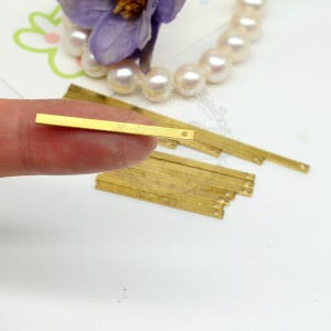May include: Close-up of several small, rectangular, gold-colored metal bars. One bar is held between fingers, showing a small hole at one end. Other bars are stacked in the background. A flower and pearls are in the background.