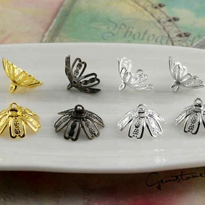 Könnte beinhalten: Eine Auswahl von acht Metall-Schmuckteilen in Gold, Schwarz und Silber. Die Teile umfassen filigrane blütenförmige Perlenkappen und offene, schmetterlingsförmige Komponenten, die auf einem weißen Teller präsentiert werden.