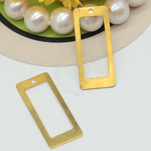 May include: Two gold-colored rectangular metal charms with a hole at the top for stringing. The charms have a hollow center and a smooth, shiny finish. The background includes pearls and a green and white design.