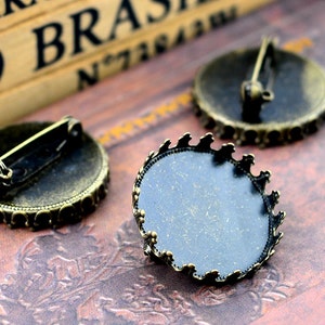 May include: Close-up of three antique bronze-toned round brooch bases. Each base has a decorative scalloped edge and a pin backing. The bases are empty, ready for embellishment. The background is a textured, reddish-brown surface.
