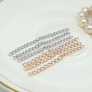 Könnte beinhalten: Nahaufnahme von Schmuckketten in Silber und Roségold. Die Ketten sind diagonal auf einer weißen Oberfläche angeordnet. Eine Perlenkette ist im Hintergrund zu sehen. Diese Ketten sind ideal für die Schmuckherstellung.