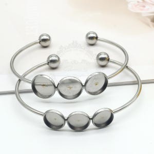 May include: Two silver-toned metal bangle bracelets. Each bracelet has a circular design with three round, recessed settings for embellishments. The bracelets have a simple, elegant design, with a small ball at each end.