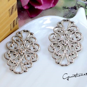 May include: Two silver filigree oval embellishments with intricate floral and scrollwork designs. The embellishments are displayed on a white surface, with the text "Gemstones2u" in the bottom right corner.