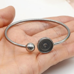 May include: A silver-colored bangle bracelet with a round ball and a circular setting. The bracelet is made of a shiny metal and is designed to hold a small item. The setting is a dark gray color.