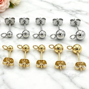 May include: A collection of ten metal stud earring findings arranged in two rows. The top row features five silver-toned ball studs with attached loops and butterfly backs. The bottom row displays five gold-toned ball studs with loops and backs.