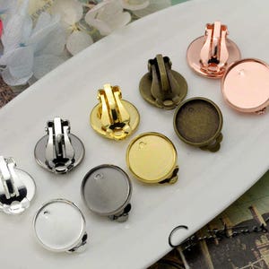 Puede incluir: Una colección de bases para pendientes de clip en varios acabados metálicos, incluyendo plata, oro, bronce y cobre. Cada base tiene una base redonda con un mecanismo de clip en la parte posterior, diseñado para sujetar pendientes. Las bases se muestran en una fuente blanca de forma ovalada.