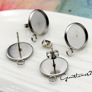 Puede incluir: Dos pares de bases de pendientes de color plateado con un engaste redondo y un anillo de salto. Las bases de pendientes están sobre una superficie blanca.