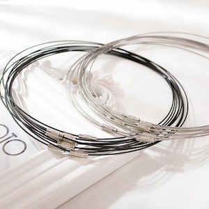 Może przedstawiać: Zestaw srebrnych i czarnych metalowych łańcuszków do naszyjników. Łańcuszki wykonane są z cienkiego drutu i mają prosty, minimalistyczny design. Łańcuszki są ułożone w kółko, ze srebrnymi łańcuszkami na czarnych łańcuszkach.