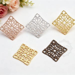 Op de afbeelding: Vijf vierkante filigraan metalen versieringen in verschillende kleuren: roségoud, zilver, goud en brons. Elk stuk heeft een open ontwerp met een centraal cirkelvormig element en stralende patronen, voor sieraden of knutselen.