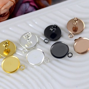 May include: Six earring settings with different colored metal finishes. The settings are round and have a flat surface for attaching a cabochon. The settings are silver, gold, black, and rose gold.