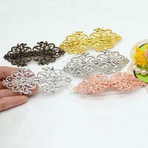 Könnte beinhalten: Fünf verzierte Haarspangen aus Metall in verschiedenen Farben: Bronze, Gold, Silber und Roségold. Jede Spange hat ein filigranes, offenes Design. Die Spangen werden auf einer weißen Oberfläche präsentiert.